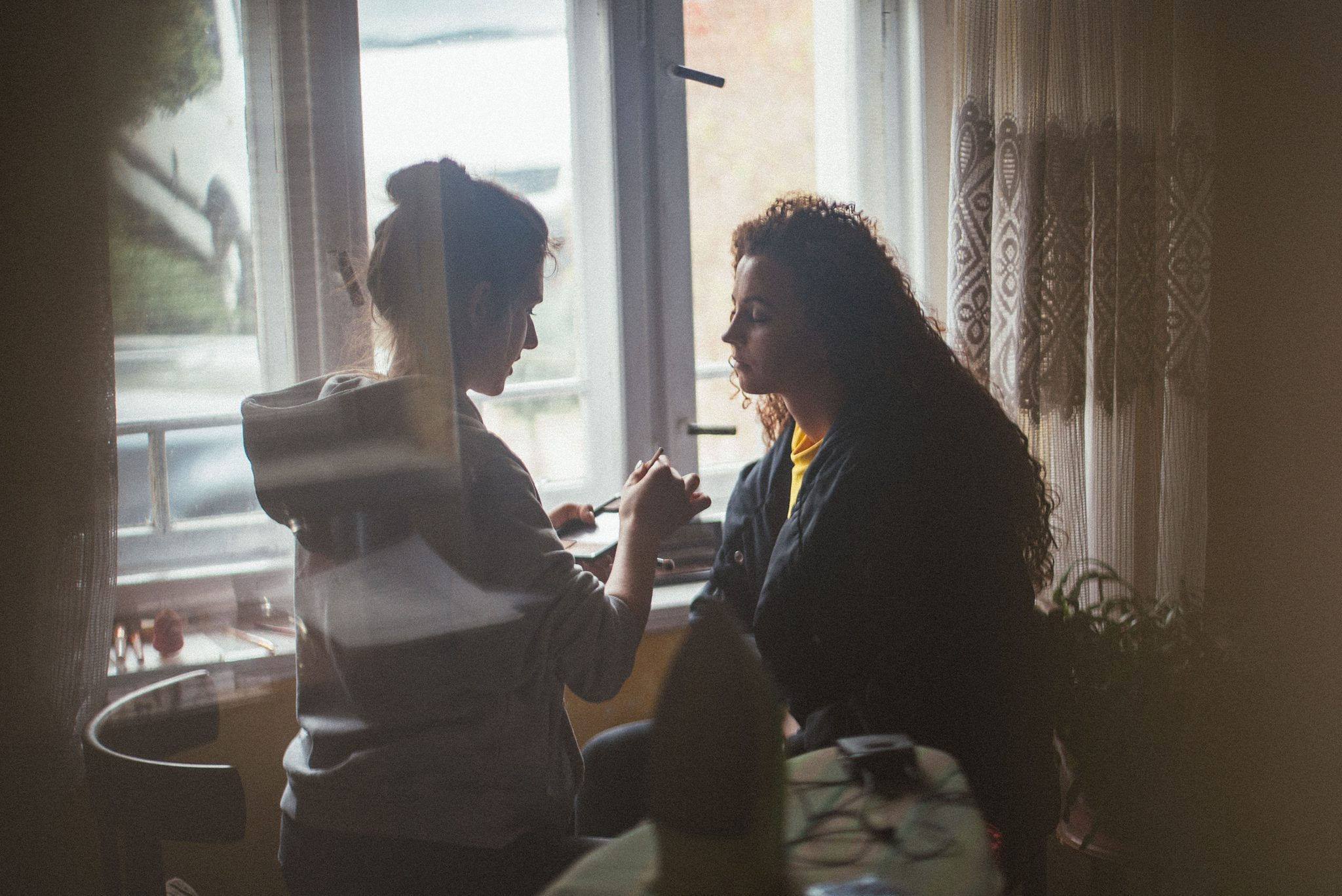 Kobieta nakłada makijaż drugiej kobiecie w pobliżu okna z zasłonami, w tle rozmazane przedmioty.