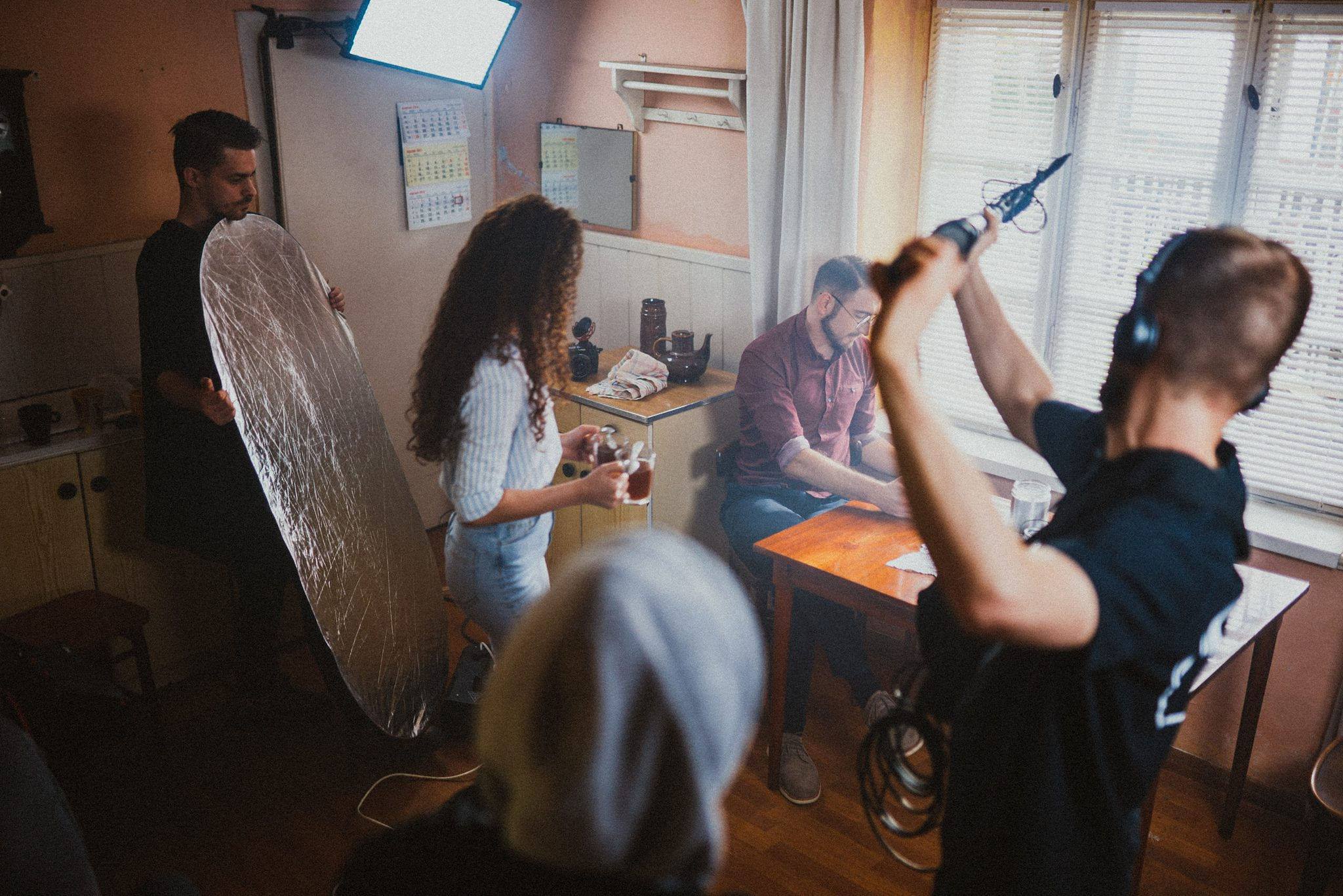 Ekipa filmowa podczas kręcenia sceny w mieszkaniu we Wrocławiu: operator z mikrofonem, oświetleniowiec z blendą, aktorka z rekwizytami i aktor przy stole.