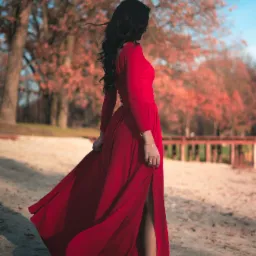 Kobieta w długiej, czerwonej sukni z rozcięciem, stojąca na piasku z drzewami w tle, włosy falują na wietrze.