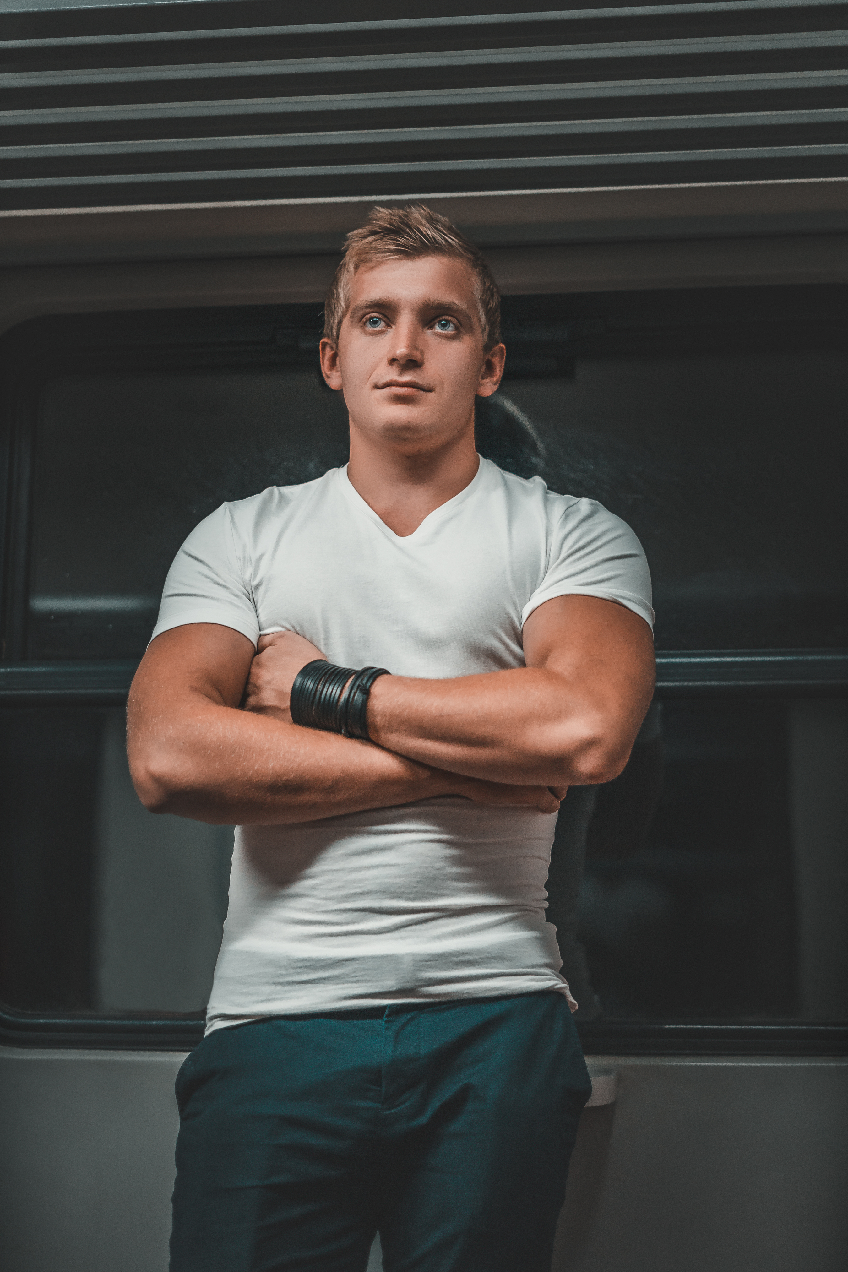 Młody mężczyzna w białej koszulce i ciemnych spodniach, z założonymi rękami i czarną skórzaną bransoletą na tle ciemnego wnętrza z poziomymi liniami, patrzy w górę.