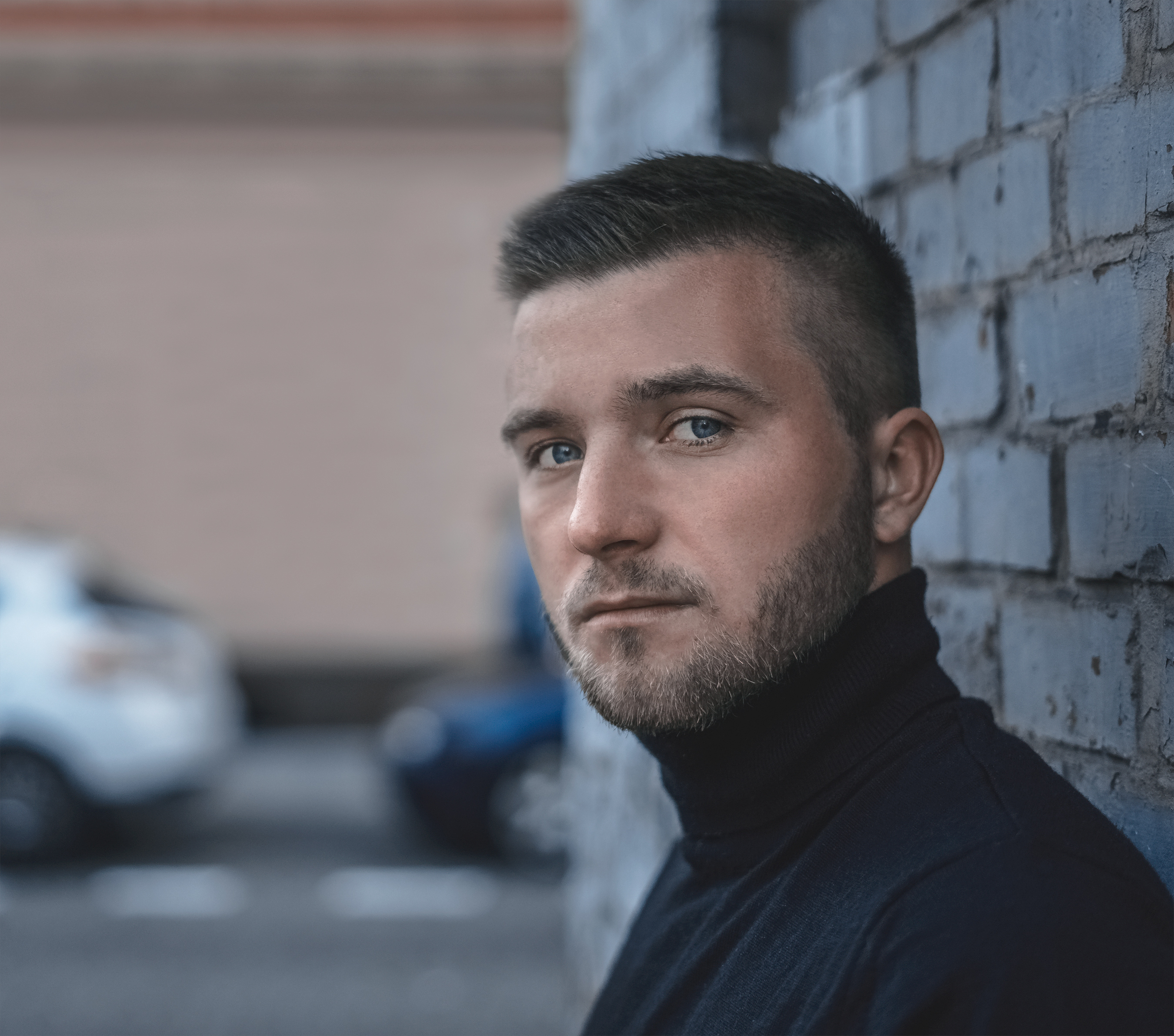 Portret młodego mężczyzny w czarnym golfie, opierającego się o szarą, ceglaną ścianę na tle rozmytych samochodów i budynku.