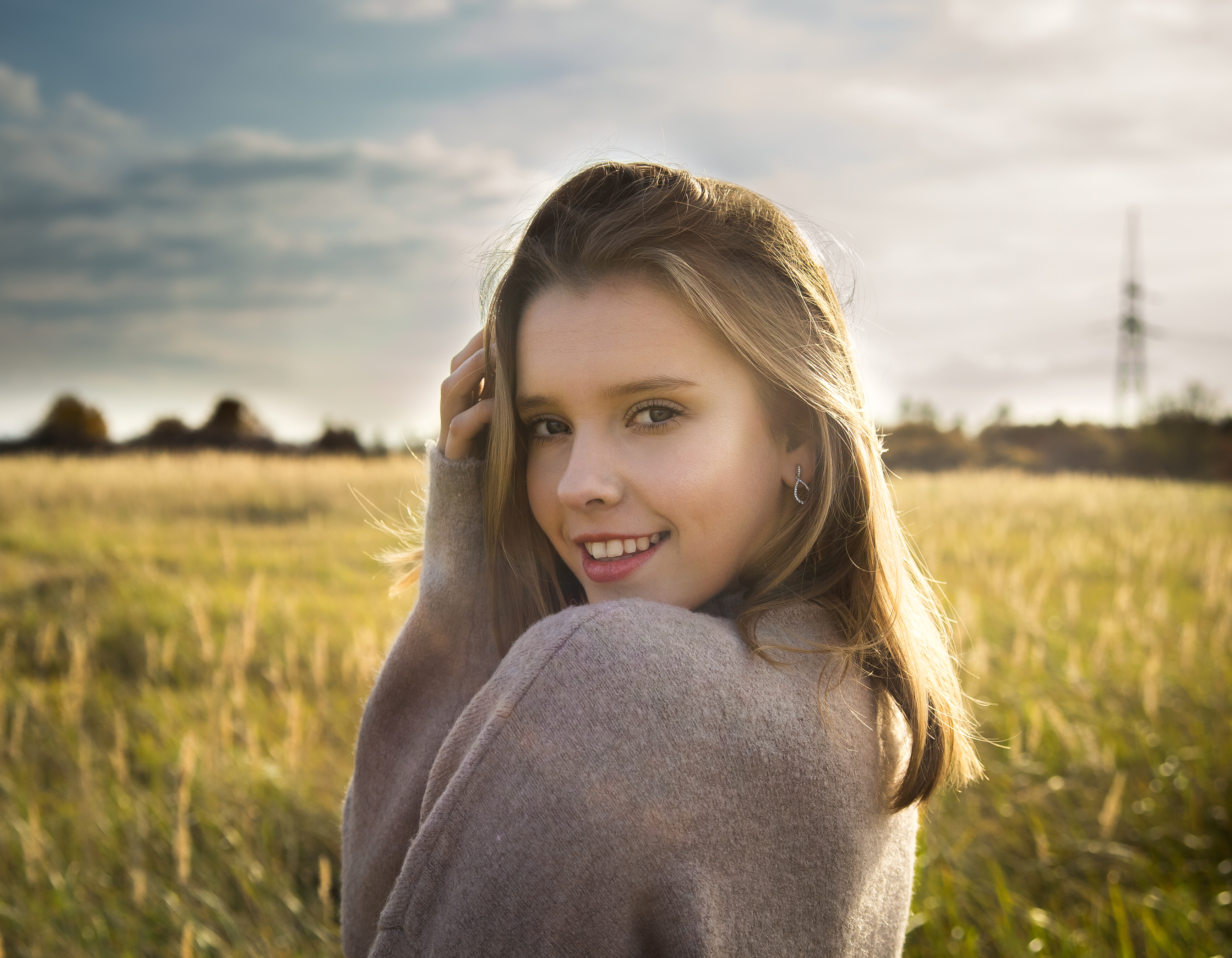 Portret młodej kobiety z uśmiechem, w beżowym swetrze, na tle pola z wysoką trawą i słupów energetycznych w oddali, z ręką we włosach i delikatnymi kolczykami.