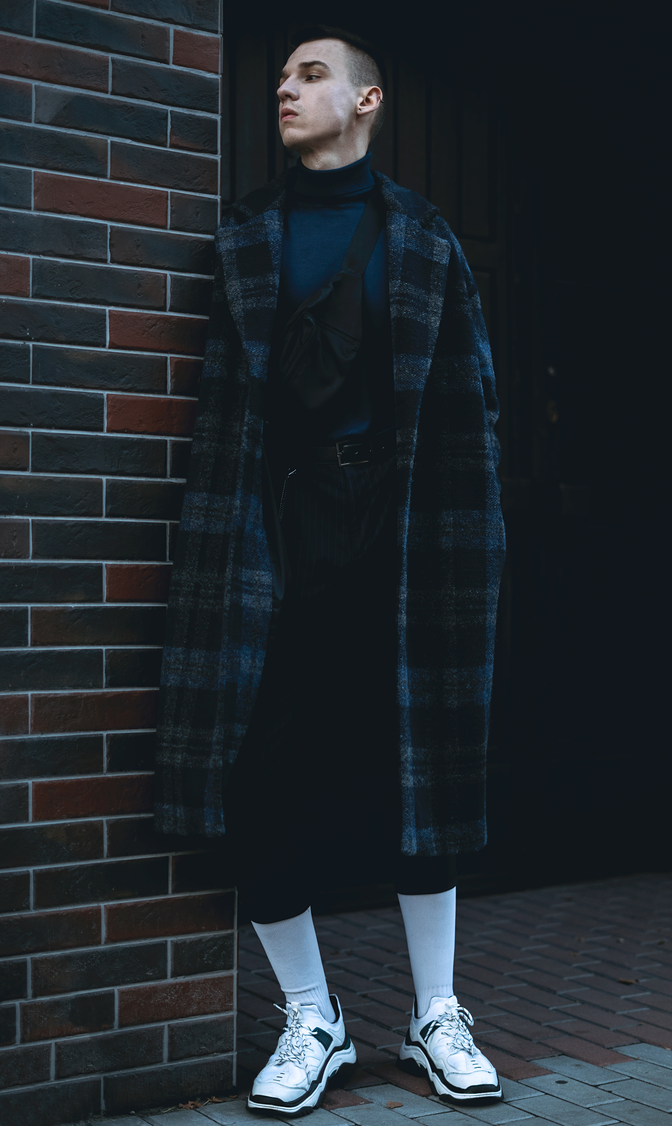 Młody mężczyzna w płaszczu w kratę, golfie i białych skarpetach pozuje na tle ceglanej ściany.