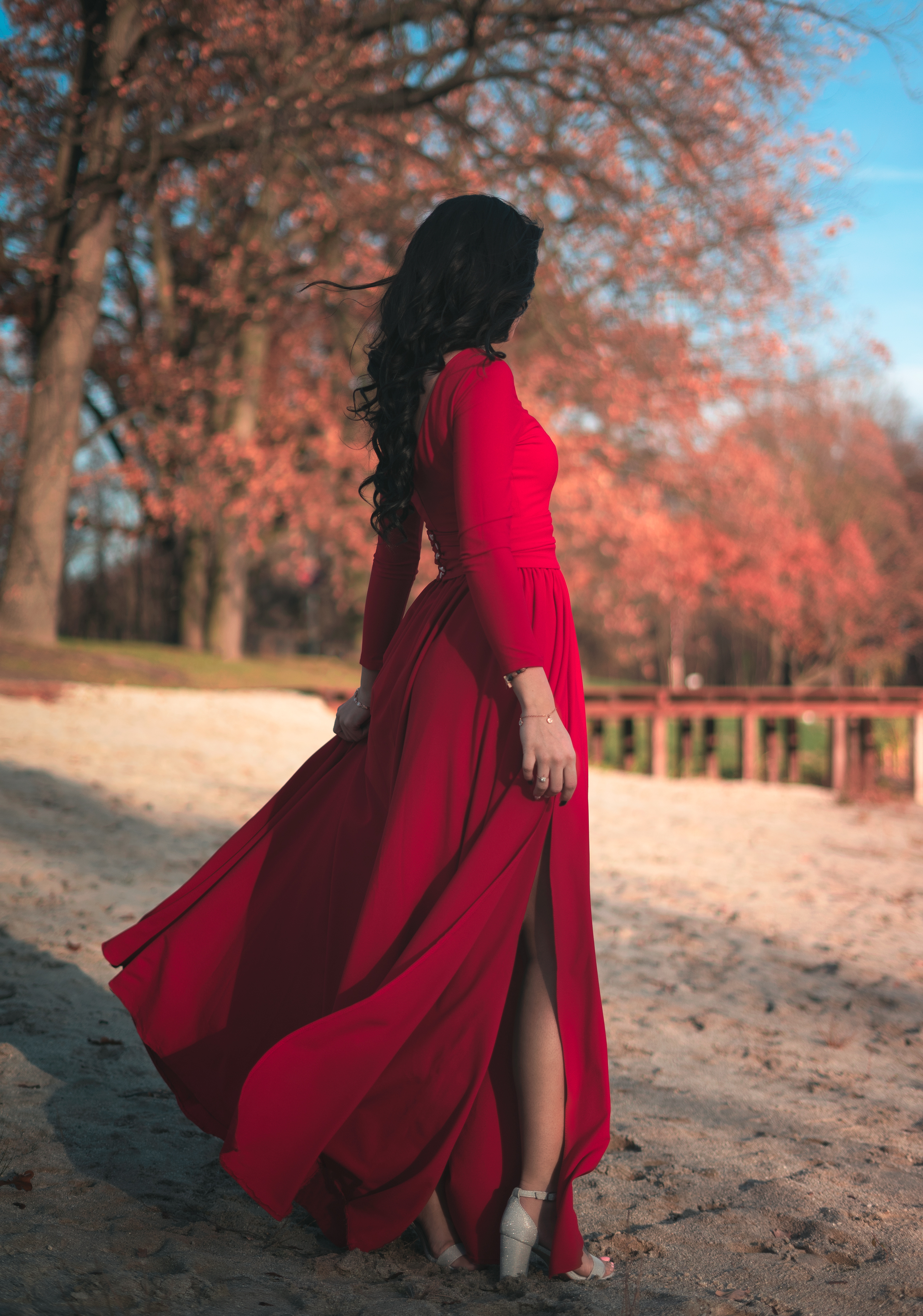 Kobieta w długiej, czerwonej sukni z rozcięciem, stojąca na piasku z drzewami w tle, włosy falują na wietrze.