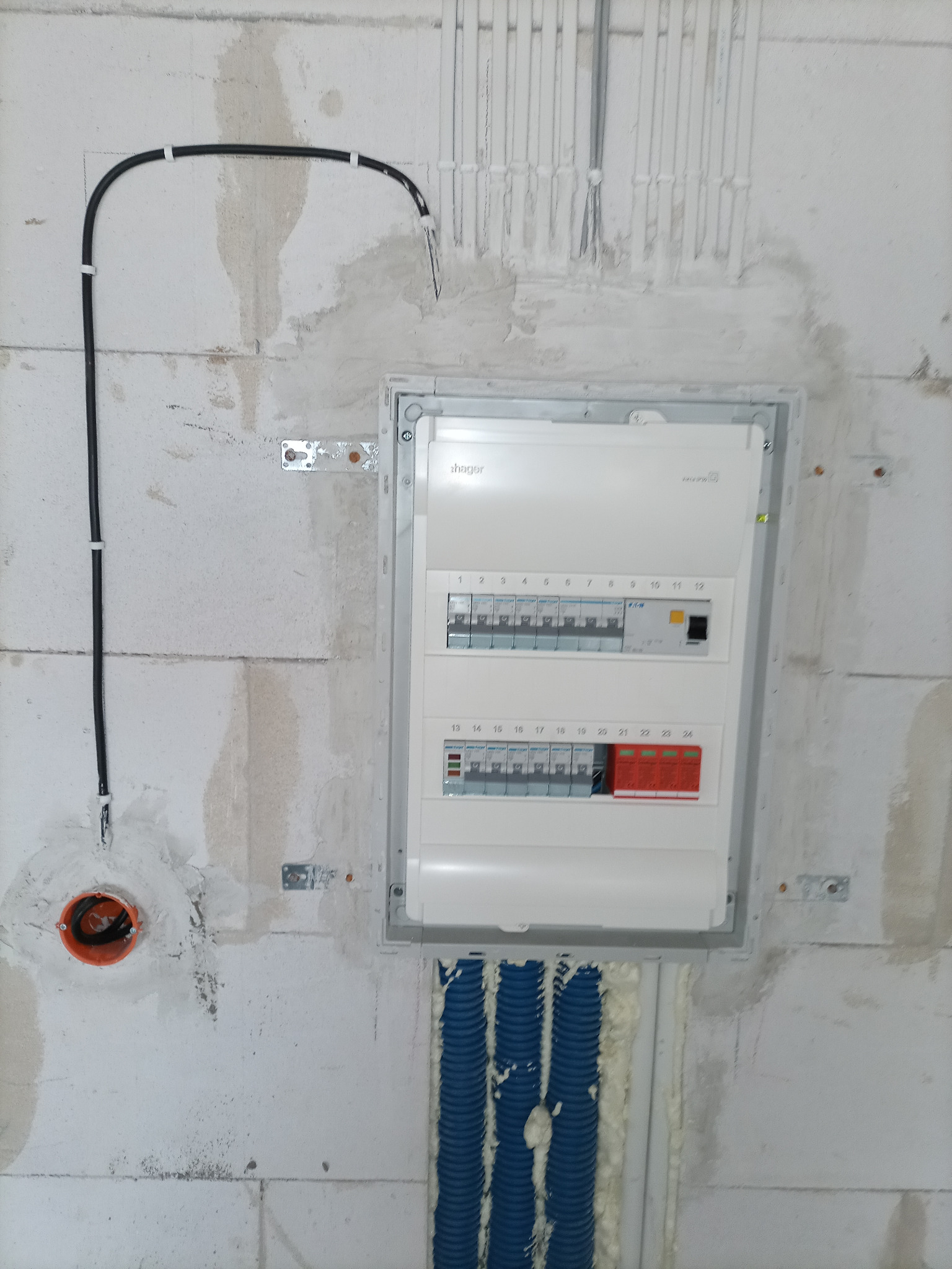 Nowa, biała skrzynka bezpiecznikowa marki Hager zamontowana na surowej, otynkowanej ścianie z betonu komórkowego, z widocznymi niebieskimi rurami karbowanymi i czarnym kablem elektrycznym w peszlu.