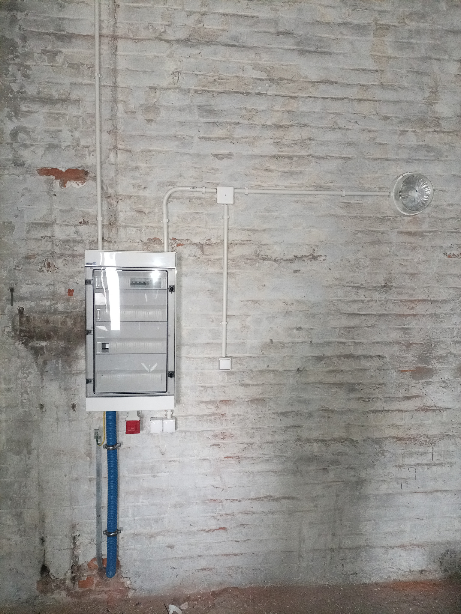 Szara, ceglana ściana z zamontowaną, białą skrzynką bezpiecznikową z przezroczystymi drzwiczkami, poprowadzone białe kable w peszlu oraz niebieska rura ochronna z uchwytami.