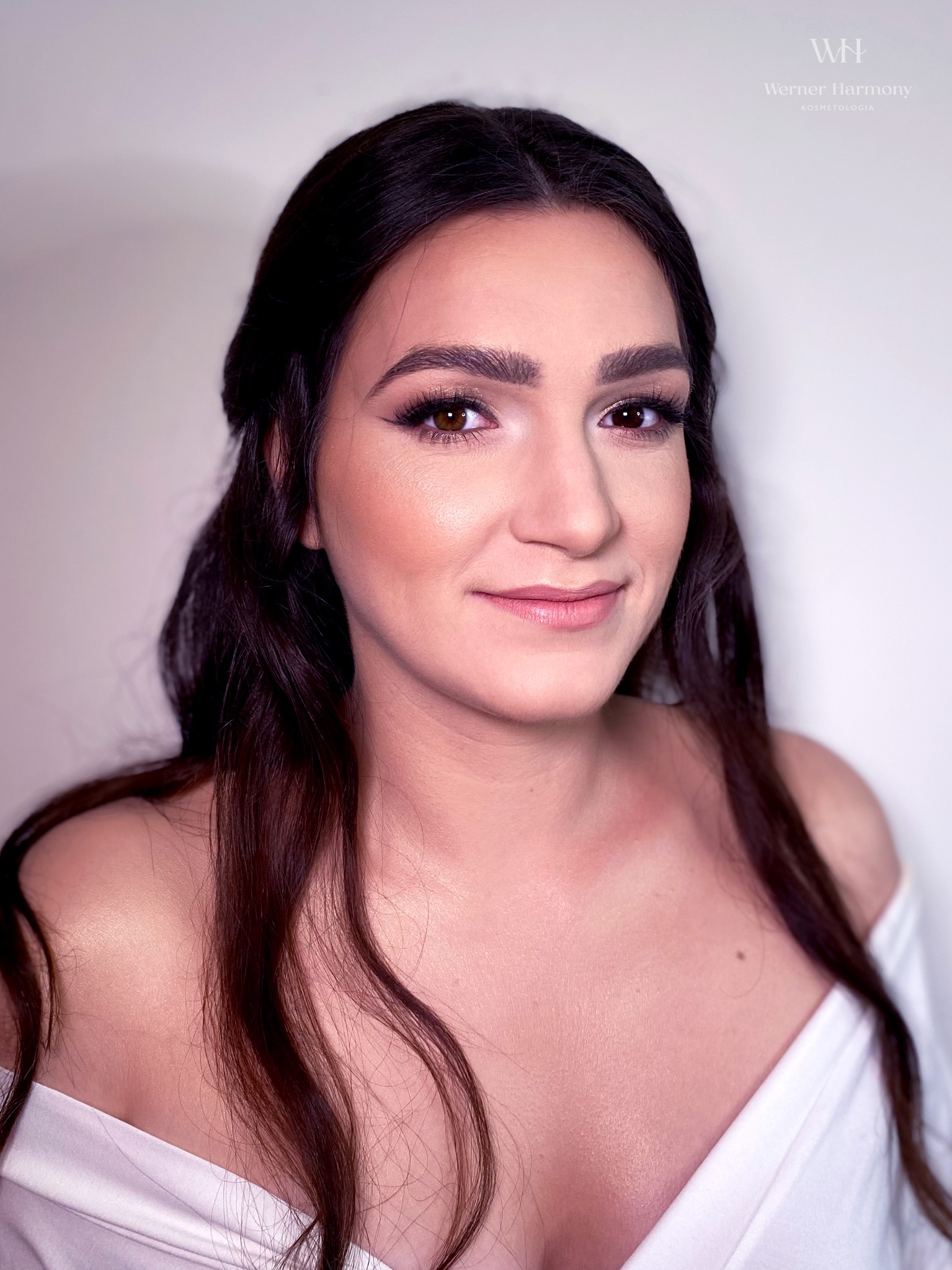 Portret uśmiechniętej brunetki z delikatnym konturowaniem twarzy, podkreślonymi oczami i błyszczącymi ustami w odcieniu nude, włosy upięte w luźne fale.