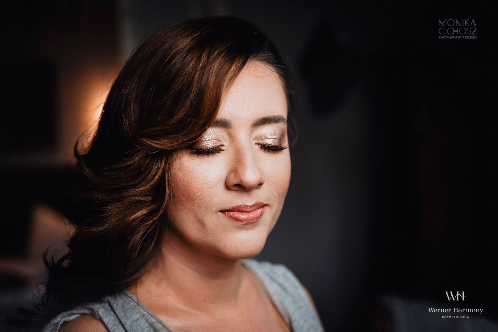 Portret kobiety z zamkniętymi oczami, prezentujący błyszczący cień do powiek w odcieniach złota i delikatny połysk na ustach, z brązowymi, falowanymi włosami opadającymi na ramię.