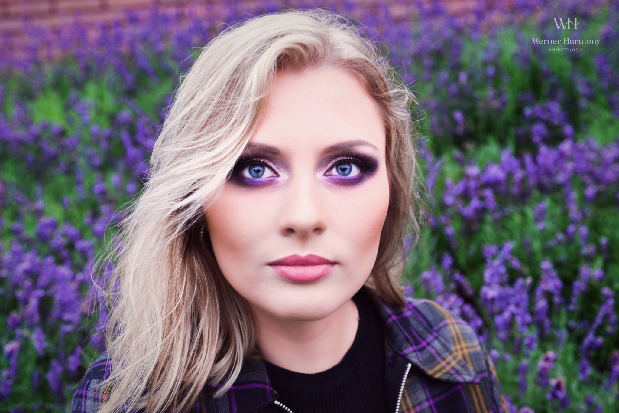 Portret kobiety z intensywnym, fioletowym cieniem na powiekach i rozświetlonym wewnętrznym kącikiem oka, na tle kwitnącej lawendy, w rozpiętej koszuli w kratę. Widoczne logo firmy Werner Harmony...