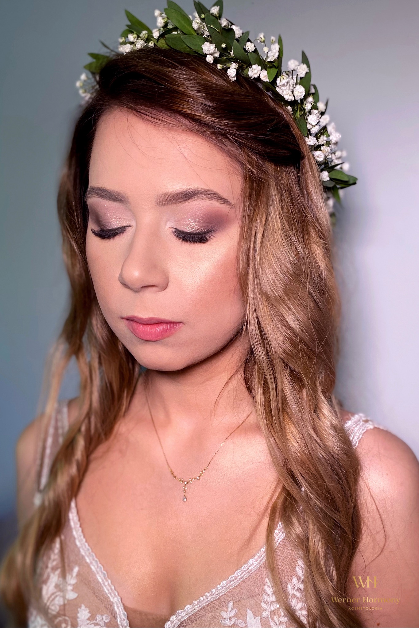 Panna młoda z zamkniętymi oczami, wiankiem z białych kwiatów i zielonych liści we włosach, delikatny róż na powiekach i ustach, złoty łańcuszek z drobnymi kamieniami na szyi, koronkowy dekolt sukni.