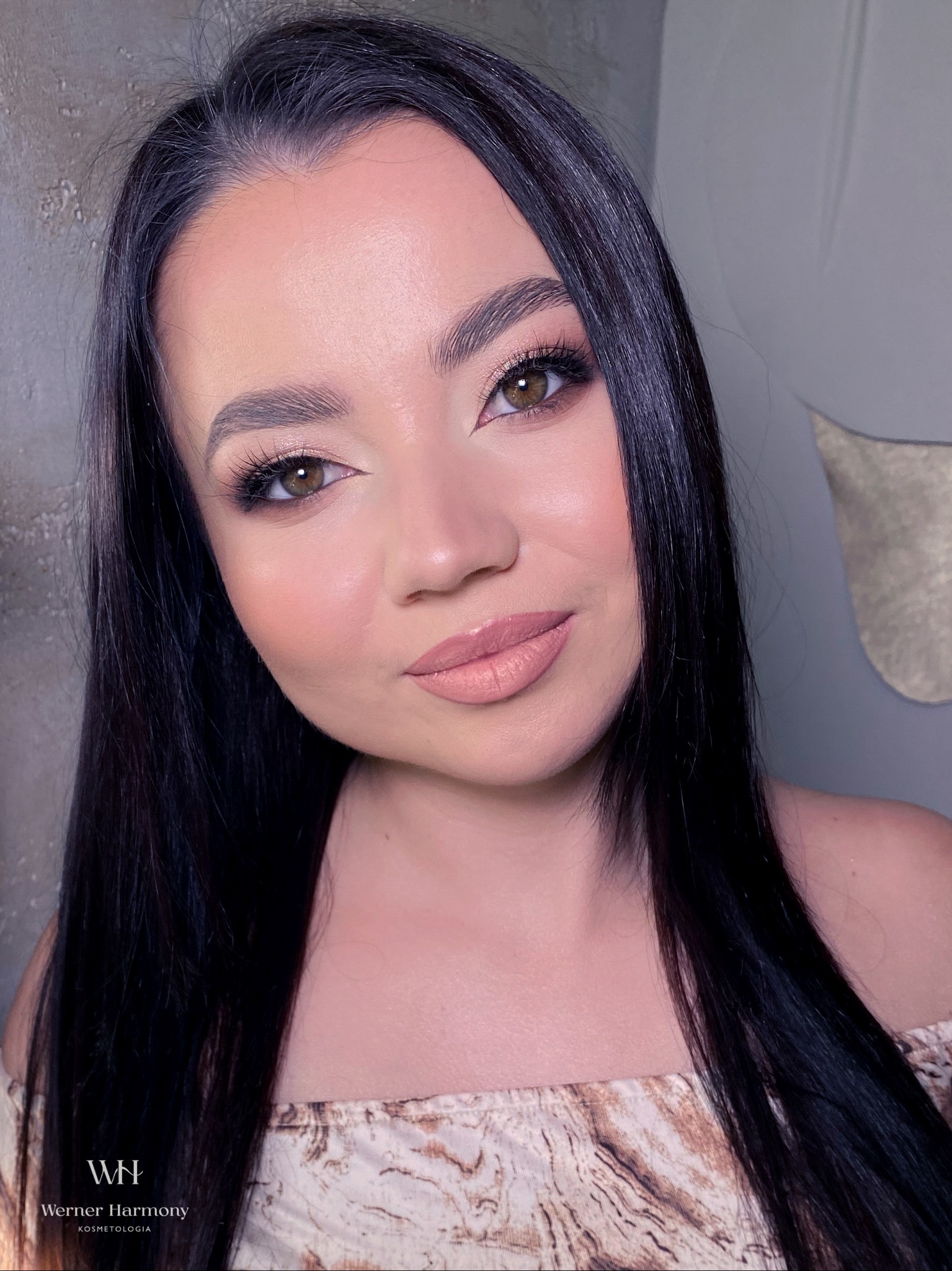 Portret kobiety z delikatnym, rozświetlonym upiększeniem twarzy, podkreślonymi brwiami i długimi, czarnymi włosami na tle szarej ściany.