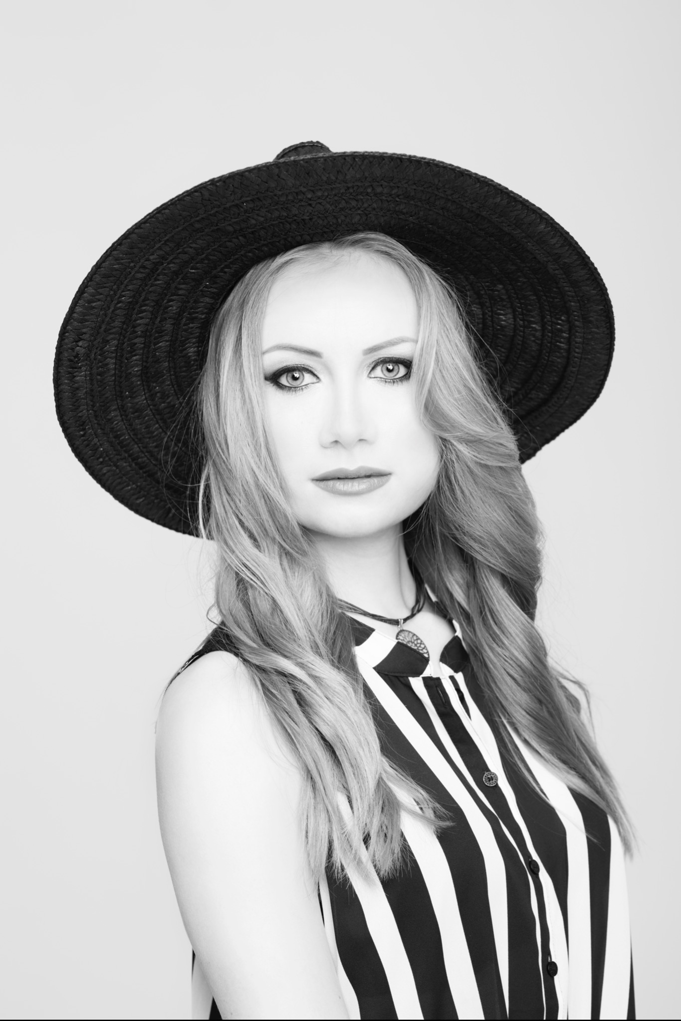 Portret kobiety w czarno-białej tonacji, noszącej kapelusz z szerokim rondem i bluzkę w paski, z delikatnym podkreśleniem oczu i ust.