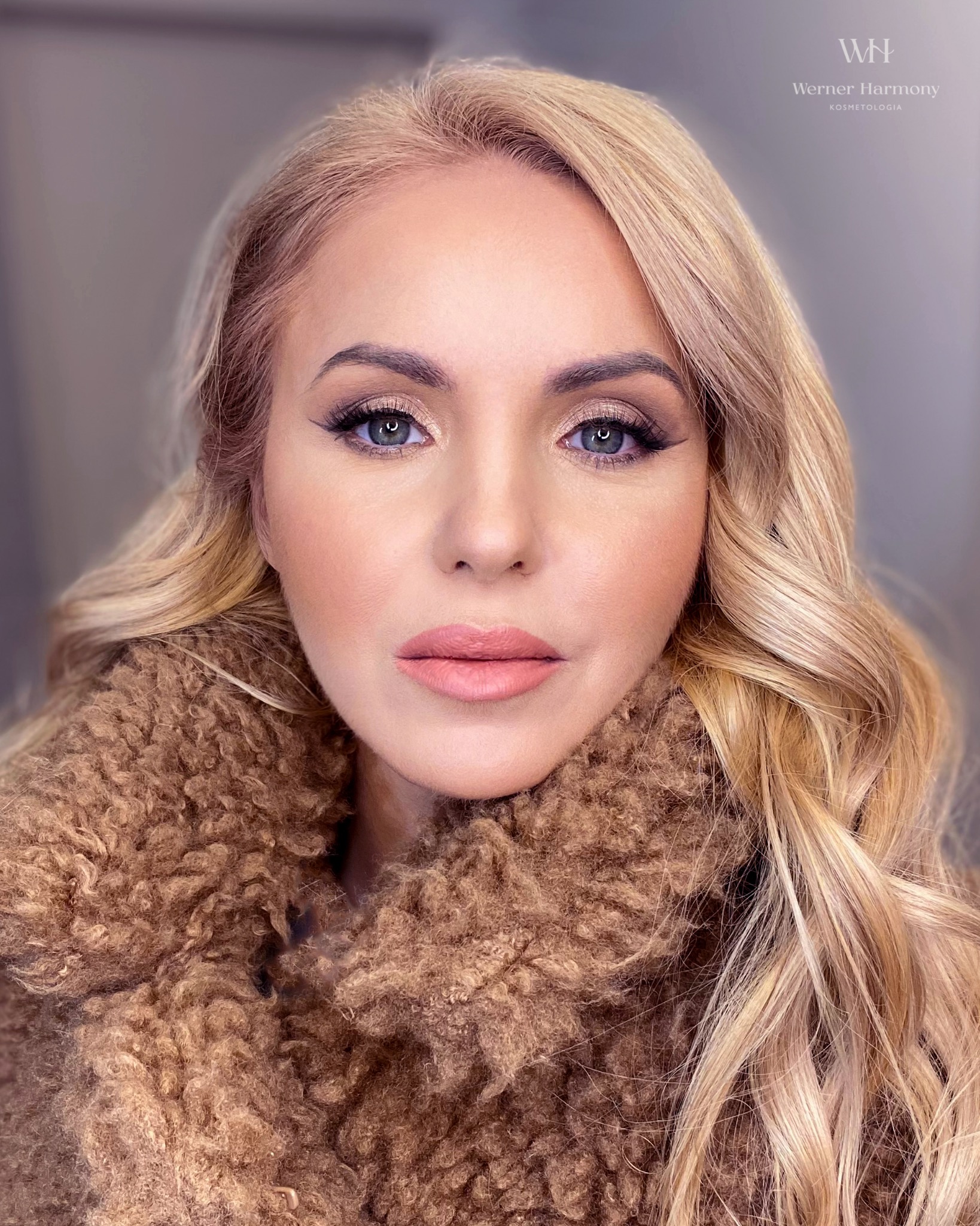 Portret kobiety z profesjonalnym, subtelnym podkreśleniem oczu eyelinerem i brązowymi cieniami, delikatnym różem na policzkach i naturalnymi ustami w otoczeniu blond fal i brązowego futra.