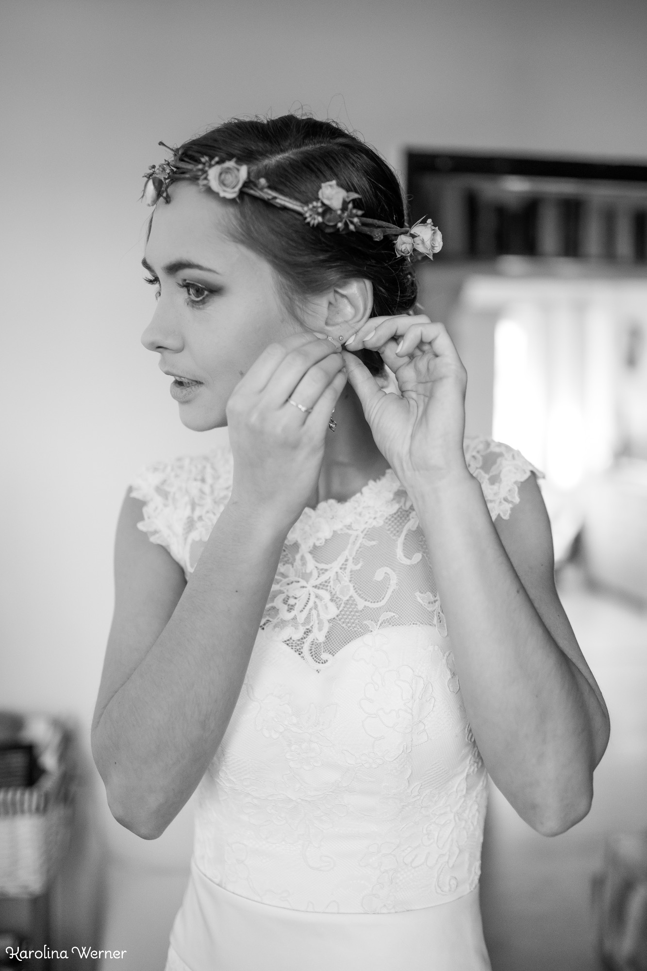 Panna młoda w koronkowej sukni i wianku z kwiatów zakłada kolczyki, profil, czarno-białe zdjęcie.