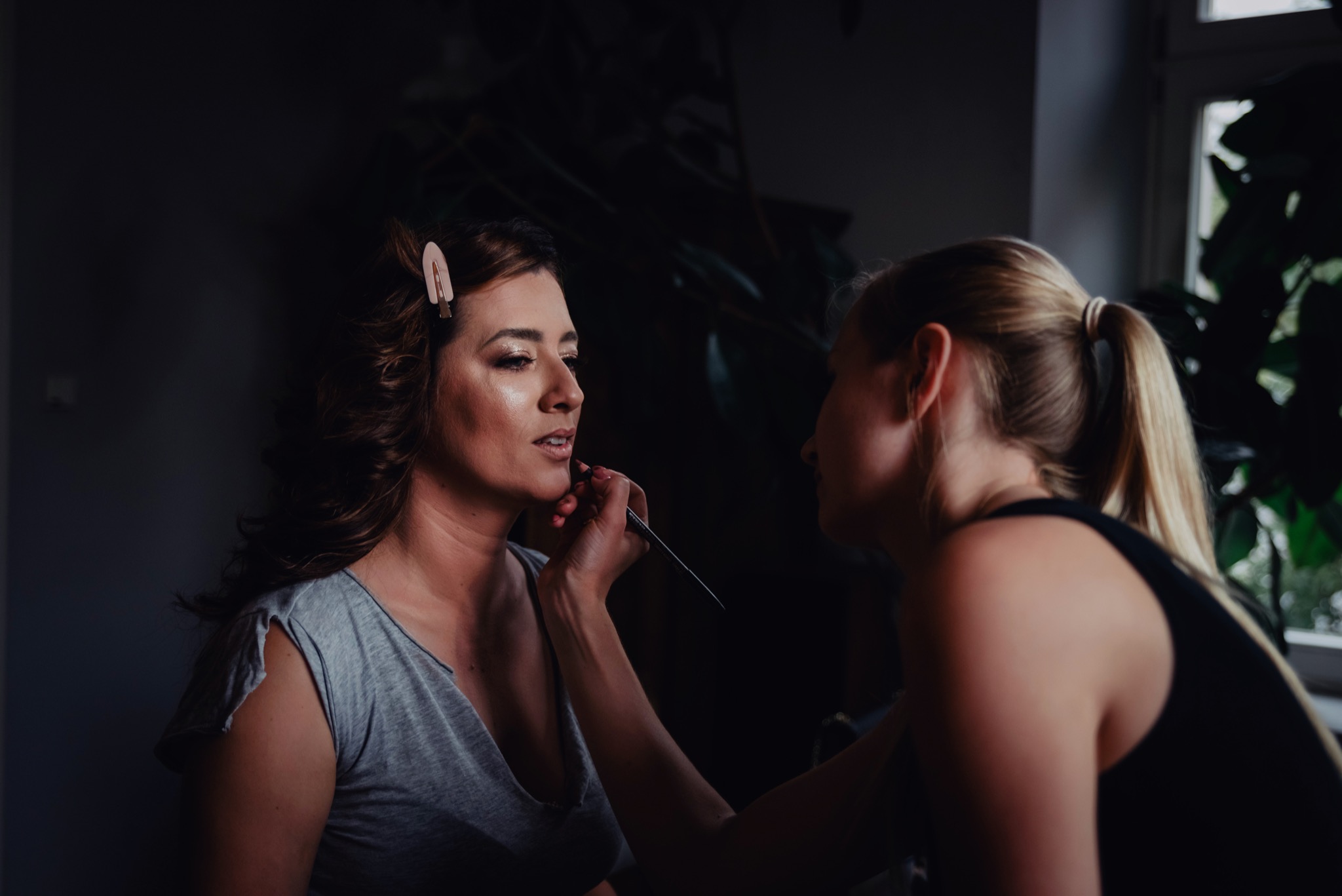 Makijażystka precyzyjnie nakłada szminkę pędzelkiem na usta klientki, widoczna spinka we włosach modelki i ciemne tło.
