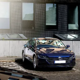 Niebieski VW Arteon zaparkowany na brukowanym parkingu przed nowoczesnym budynkiem z szarymi oknami i betonową ścianą.