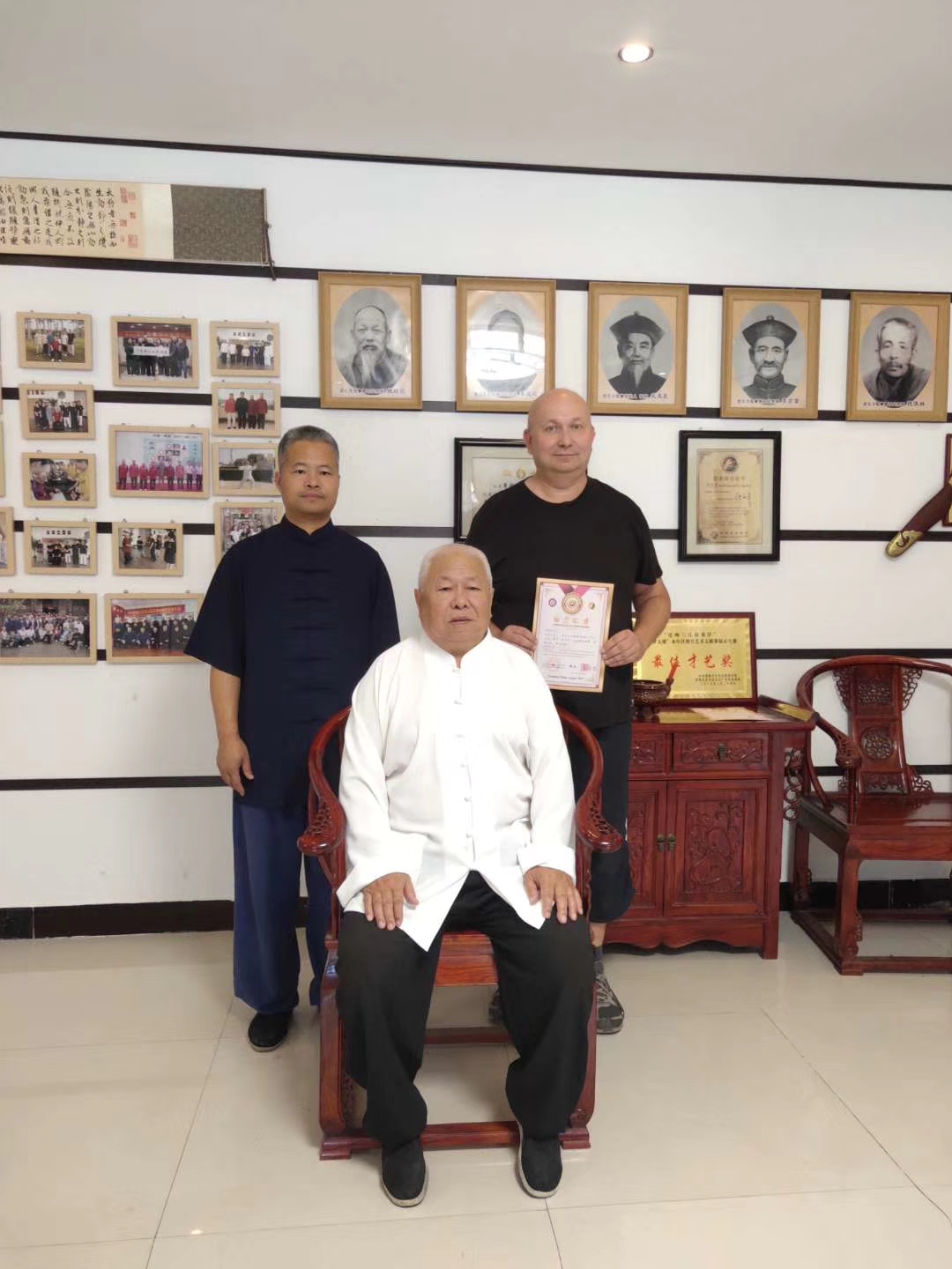 Trzy osoby pozują do zdjęcia w pomieszczeniu z portretami na ścianie; mężczyzna na środku siedzi na zdobionym krześle i trzyma certyfikat, a za nim stoi mężczyzna z europejskimi rysami twarzy.