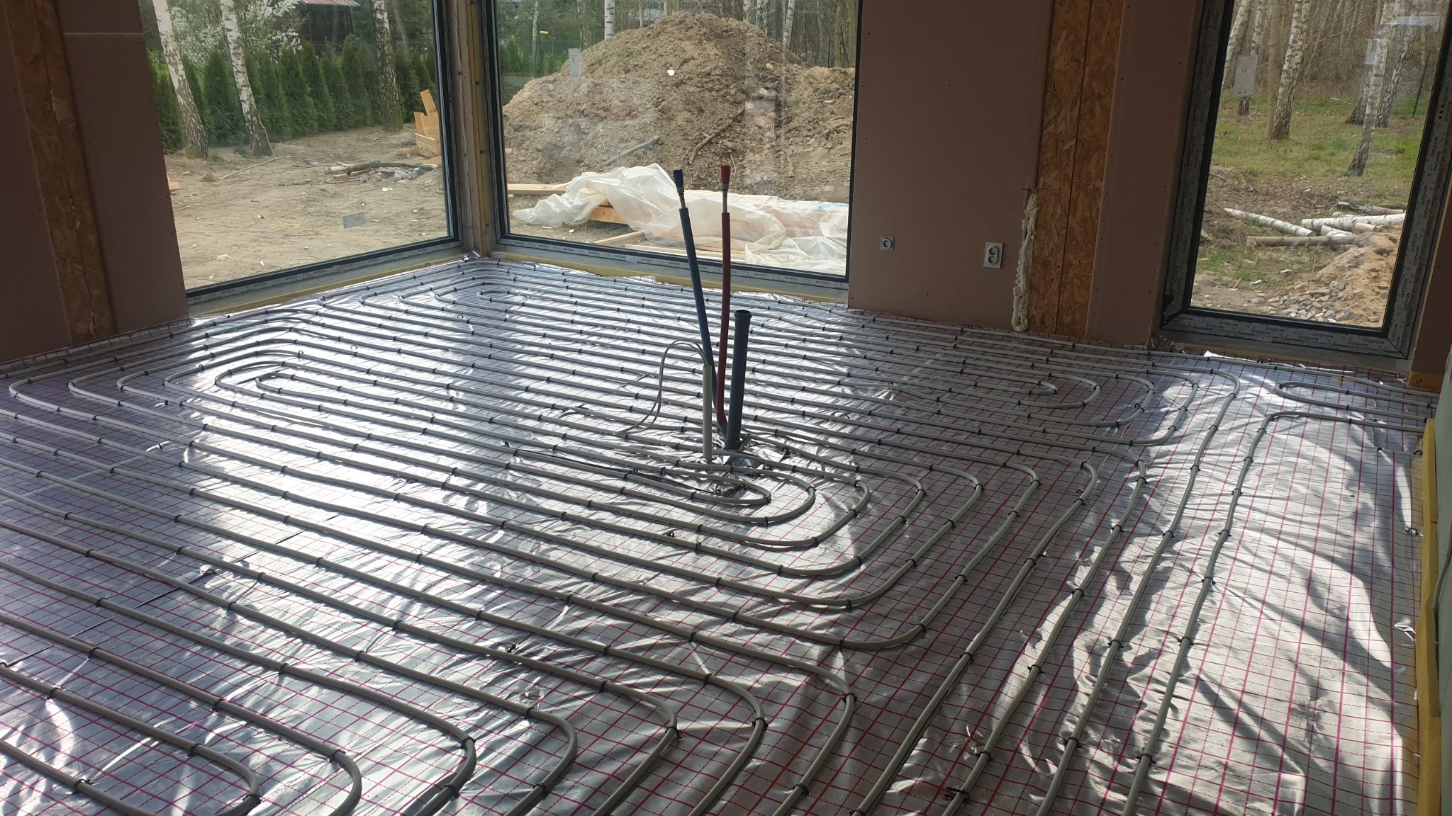 Świeżo zainstalowane rury ogrzewania podłogowego na folii z siatką, wystające rury pionowe, surowy stan wykończenia wnętrza, duże okno z widokiem na plac budowy.