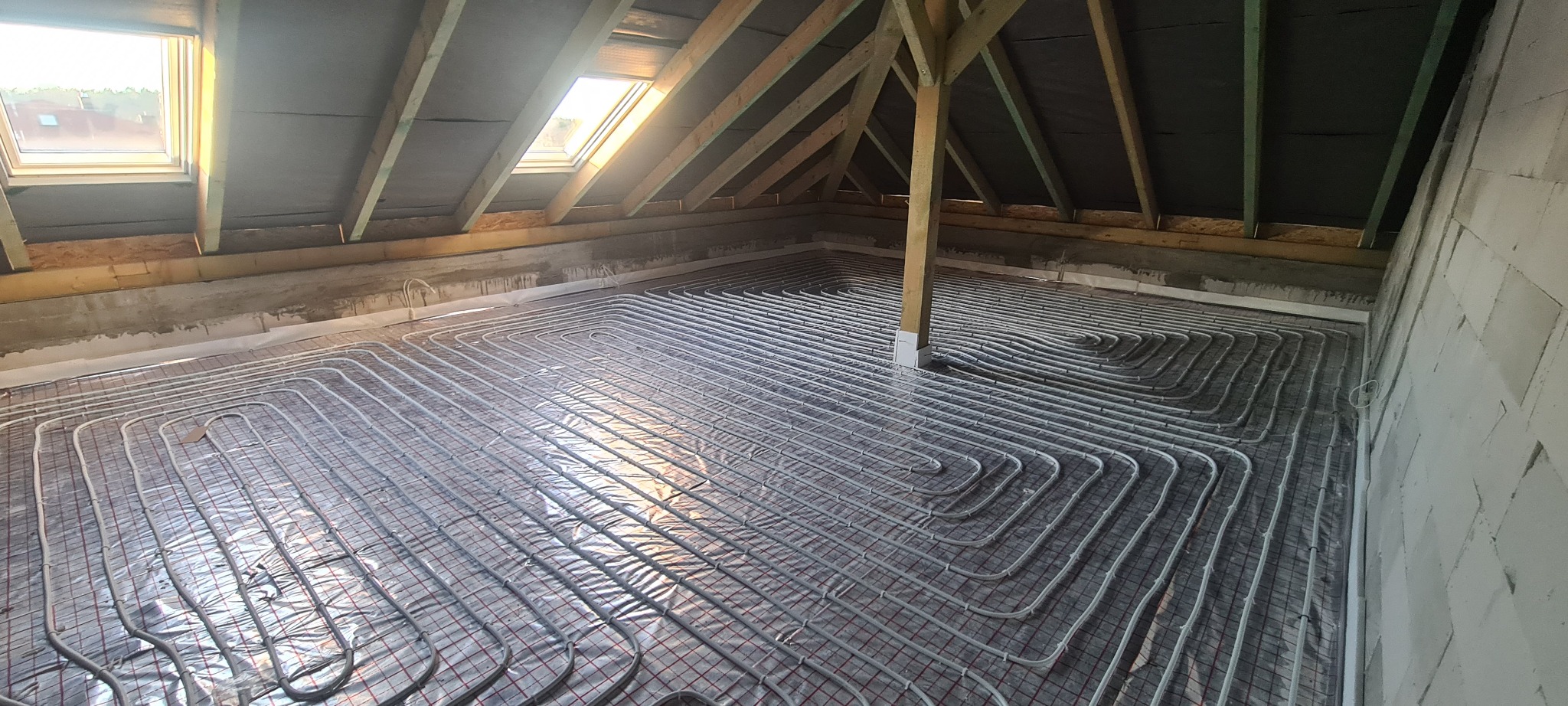 Montaż ogrzewania podłogowego w surowym wnętrzu poddasza: widok na rozłożone rury grzewcze na siatce, z oknami dachowymi i konstrukcją drewnianą w tle.