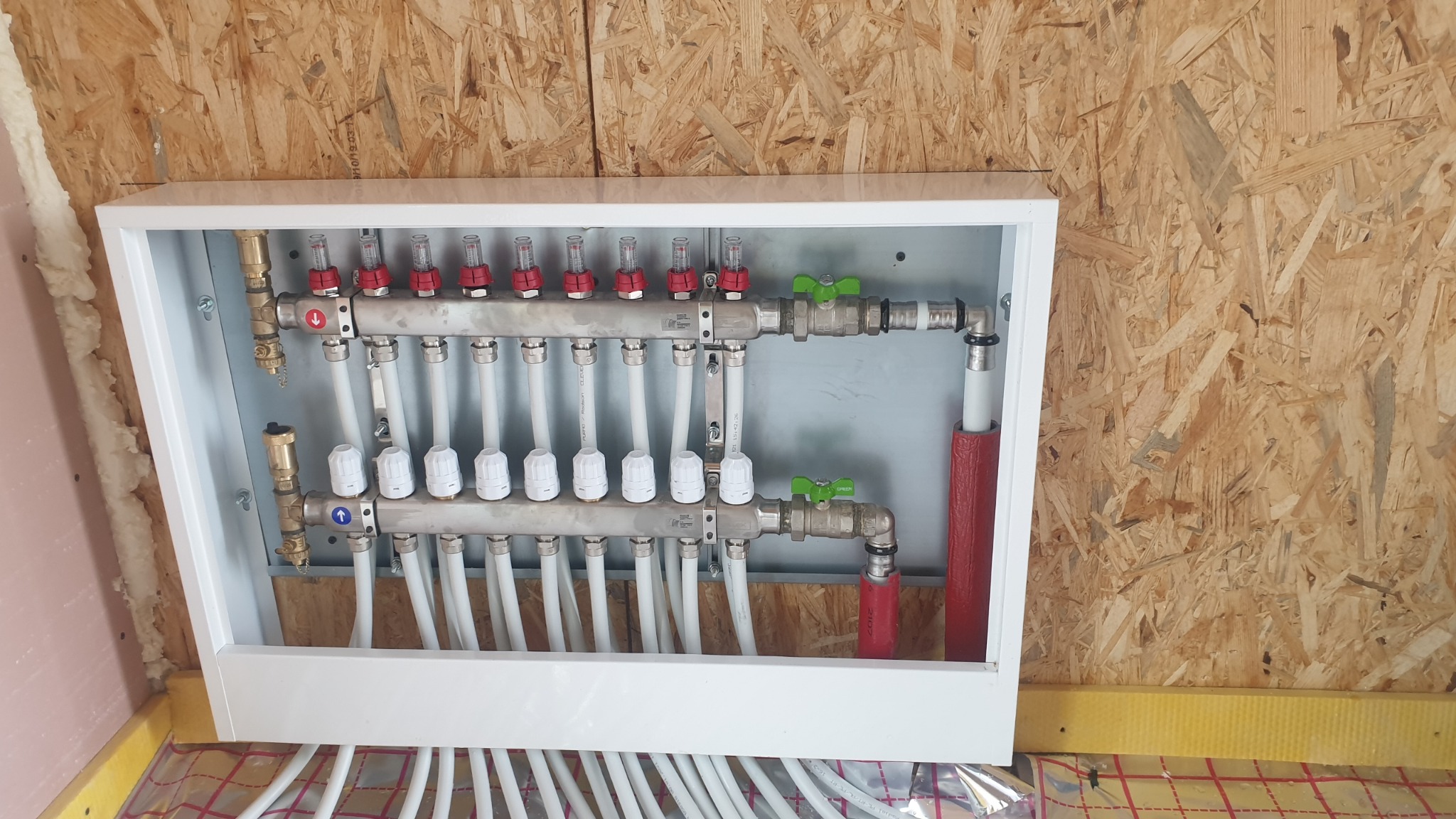 Rozdzielacz ogrzewania podłogowego w białej obudowie, zamontowany w domu w Tychach, widoczne liczne rury i zawory z elementami czerwonymi i zielonymi.