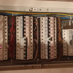 Otwarta skrzynka elektryczna z widocznymi bezpiecznikami, przewodami w kolorach niebieskim, żółtym i czerwonym oraz aparaturą modułową, z dwoma diodami sygnalizacyjnymi świecącymi na zielono...