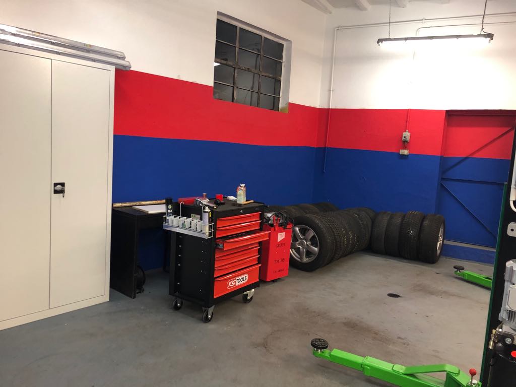 Wnętrze warsztatu samochodowego z oponami ułożonymi pod ścianą, szafką narzędziową KS Tools na kółkach i podnośnikiem samochodowym z zielonymi ramionami na szarej posadzce. Ściany pomalowane w pasy...