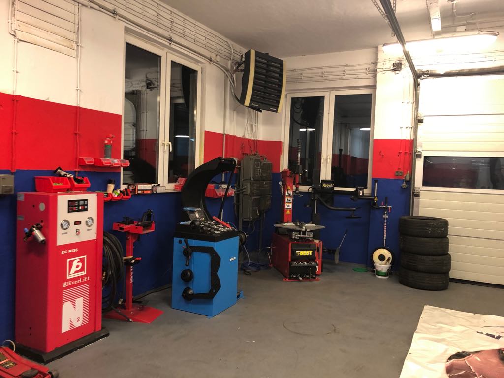 Wnętrze warsztatu wulkanizacyjnego z urządzeniami do wyważania kół i montażu opon, ściany pomalowane w kolorach czerwonym i niebieskim, stos opon w rogu.