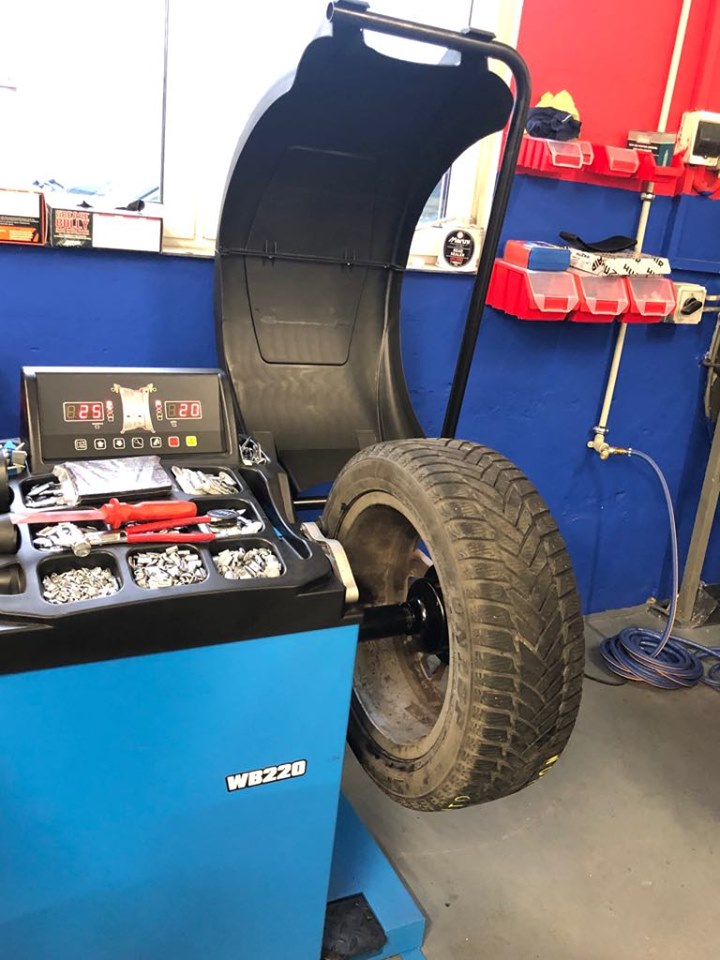 Wyważarka do kół WB220 z oponą zimową, widoczne obciążniki i narzędzia, niebieska obudowa urządzenia na tle niebiesko-czerwonej ściany warsztatu.