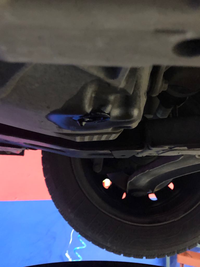 Olej silnikowy wyciekający z miski olejowej podwozia samochodu, widoczne opona i elementy zawieszenia, tło w kolorach niebieskim i czerwonym.