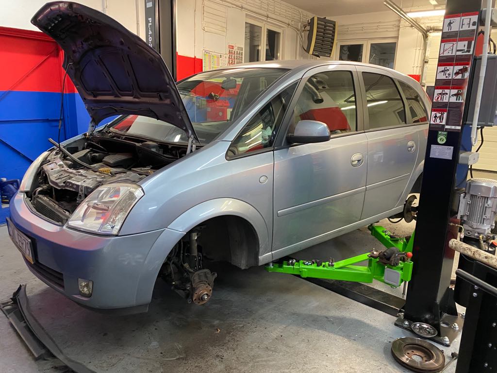 Srebrny minivan Opel Meriva bez kół, podniesiony na zielonym podnośniku w warsztacie samochodowym z otwartą maską, widoczne elementy silnika i instrukcja obsługi podnośnika na słupie.