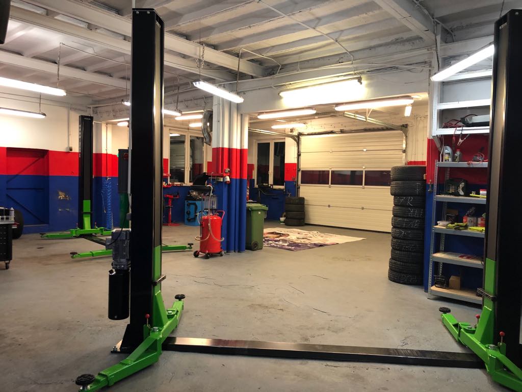 Wnętrze warsztatu samochodowego z podnośnikami, oponami i narzędziami, oświetlone lampami sufitowymi. Widoczne ściany w kolorach czerwonym i niebieskim.