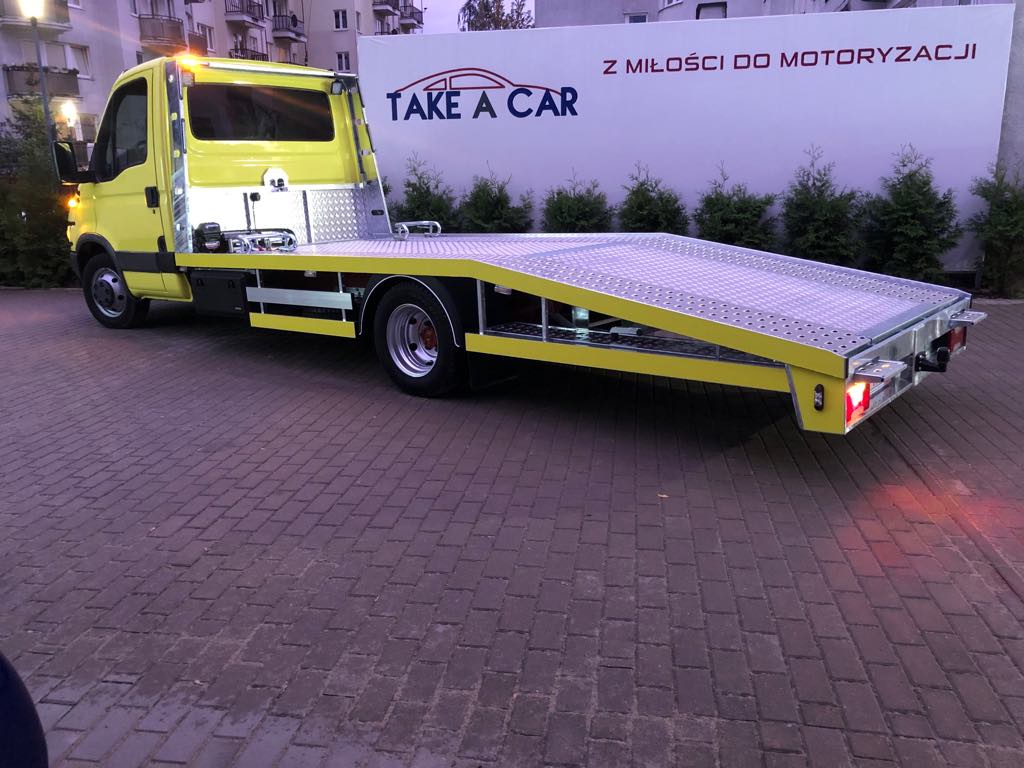 Żółta laweta z napisem 'TAKE A CAR' na tle ściany z napisem 'Z MIŁOŚCI DO MOTORYZACJI' zaparkowana na brukowanym chodniku.
