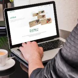 Osoba przegląda na laptopie stronę internetową z wizualizacją opakowań produktów spożywczych marki Greenergy Let's Raw, obok filiżanka kawy i roślina w doniczce.