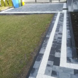 Ułożona kostka brukowa w odcieniach szarości i bieli tworząca wzór na podjeździe, z widocznym trawnikiem i kolorowymi pojemnikami na śmieci w tle.