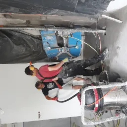 Dwóch pracowników w słuchawkach tnie ścianę piłą do betonu na szynie, podłączoną do odkurzacza przemysłowego, w pomieszczeniu zabezpieczonym folią.