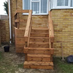 Zewnętrzne drewniane schody z podestem prowadzące do drzwi tarasowych w domu z żółtej cegły, obok widoczny fragment muru i zielona skrzynia.