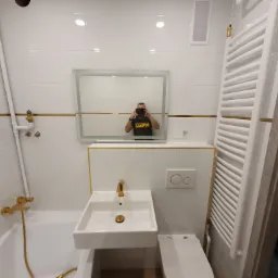 Biała łazienka z złotymi akcentami: armatura, listwy i przyciski spłuczki. W lustrze odbija się osoba robiąca zdjęcie. Widoczny grzejnik drabinkowy i umywalka.