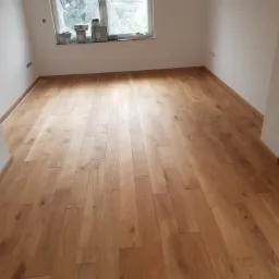 Nowo położona, jasna, drewniana podłoga panelowa w pustym, jasnym pokoju z oknem, widoczne listwy przypodłogowe.