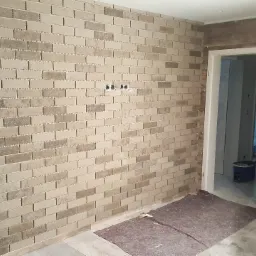 Ściana wyłożona beżowo-brązową mozaiką imitującą cegłę, widoczne puszki elektryczne, zabezpieczona podłoga i fragment ściany z paneli drewnopodobnych w trakcie montażu.