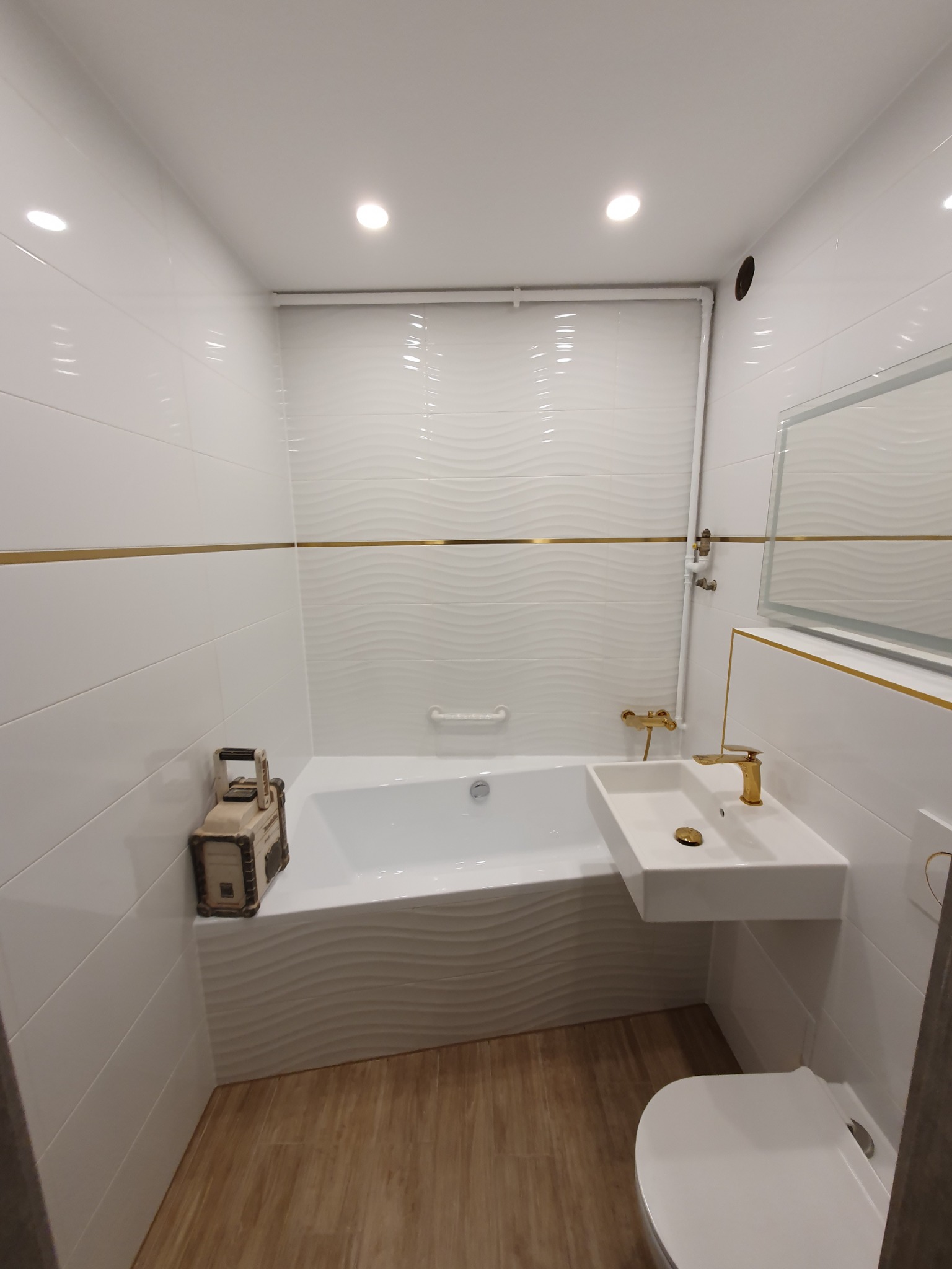 Nowoczesna łazienka z białymi płytkami o falistej strukturze, złotymi akcentami, wiszącą umywalką z baterią w kolorze złota, wanną i dekoracyjną skrzynką obok wanny.