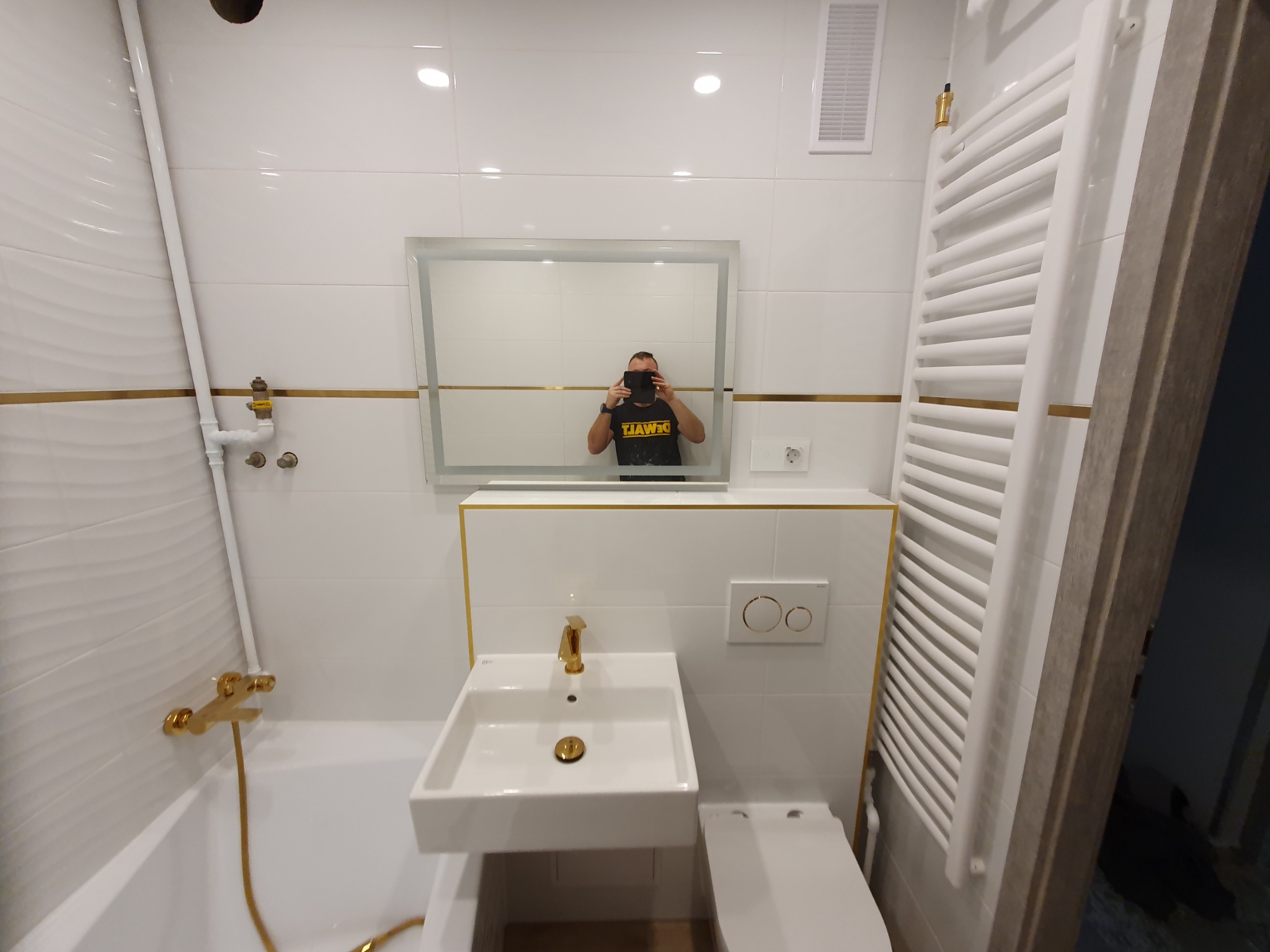 Biała łazienka z złotymi akcentami: armatura, listwy i przyciski spłuczki. W lustrze odbija się osoba robiąca zdjęcie. Widoczny grzejnik drabinkowy i umywalka.