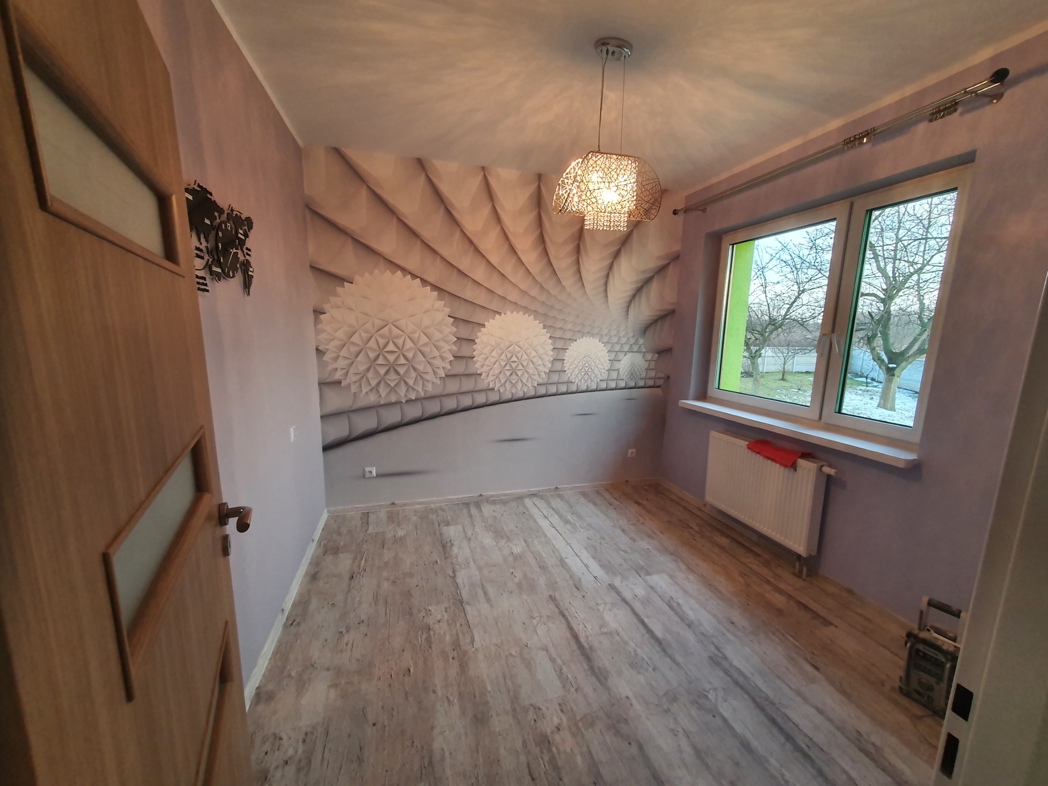 Pokój po remoncie z panelami podłogowymi imitującymi drewno, ozdobną tapetą na ścianie z motywem kulistych kwiatów i nowoczesną lampą wiszącą