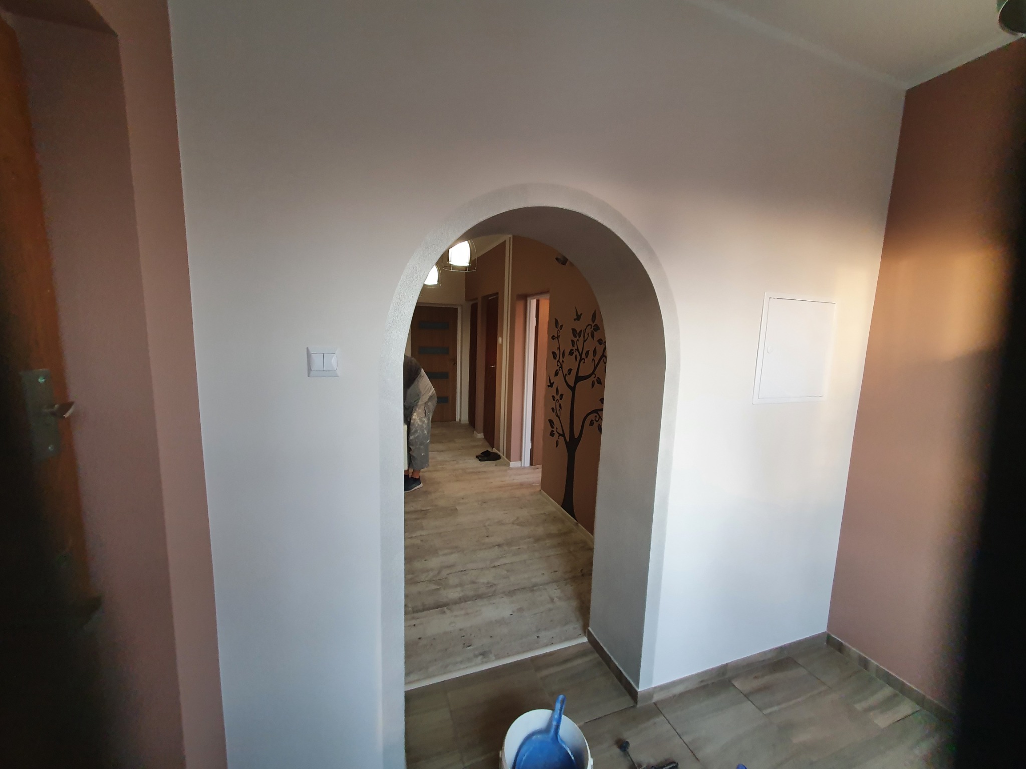 Wnętrze po malowaniu z łukowym przejściem, widoczne pomalowane ściany na biało i brązowo, jasna podłoga imitująca drewno, osoba w tle i wiadro z niebieską szufelką na pierwszym planie.