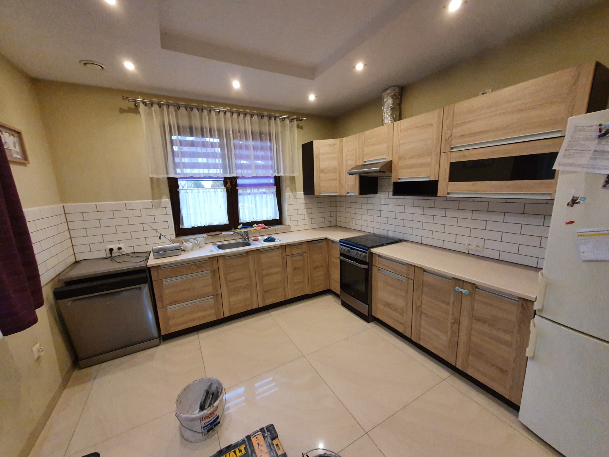 Jasna kuchnia z drewnianymi szafkami i białymi płytkami na ścianie, widoczny kubeł z narzędziami na podłodze sugerujący prace wykończeniowe.