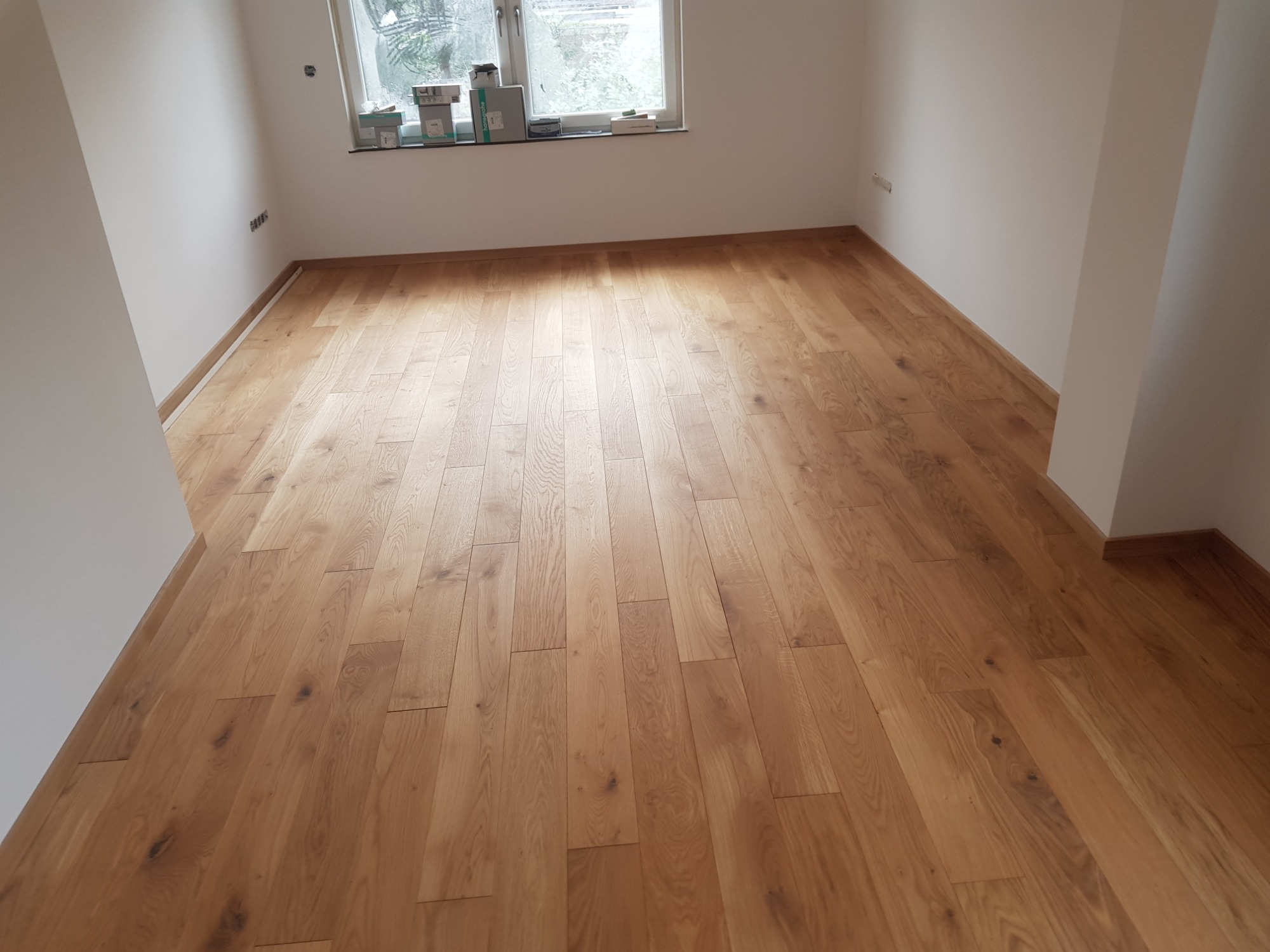 Nowo położona, jasna, drewniana podłoga panelowa w pustym, jasnym pokoju z oknem, widoczne listwy przypodłogowe.