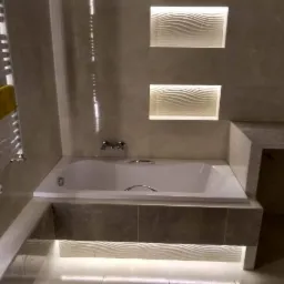 Nowoczesna łazienka w odcieniach szarości z podświetlaną wanną, dekoracyjnymi panelami ściennymi z motywem fal i grzejnikiem drabinkowym z żółtym ręcznikiem.