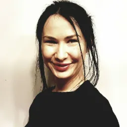 Portret uśmiechniętej kobiety z ciemnymi włosami i czerwoną szminką na tle jasnej ściany.