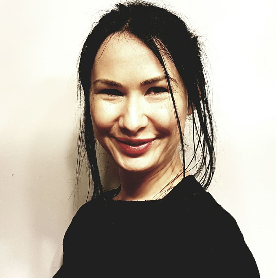 Portret uśmiechniętej kobiety z ciemnymi włosami i czerwoną szminką na tle jasnej ściany.