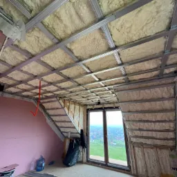 Poddasze w trakcie ocieplania: widoczne metalowe profile, wełna izolacyjna, fragment ściany z płyt kartonowo-gipsowych w kolorze różowym, okno z widokiem na zabudowania.