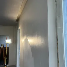 Świeżo nałożona, wilgotna gładź na ścianie w pokoju, oświetlona halogenem na statywie, widoczny fragment okna zabezpieczonego folią malarską oraz drabina w tle.